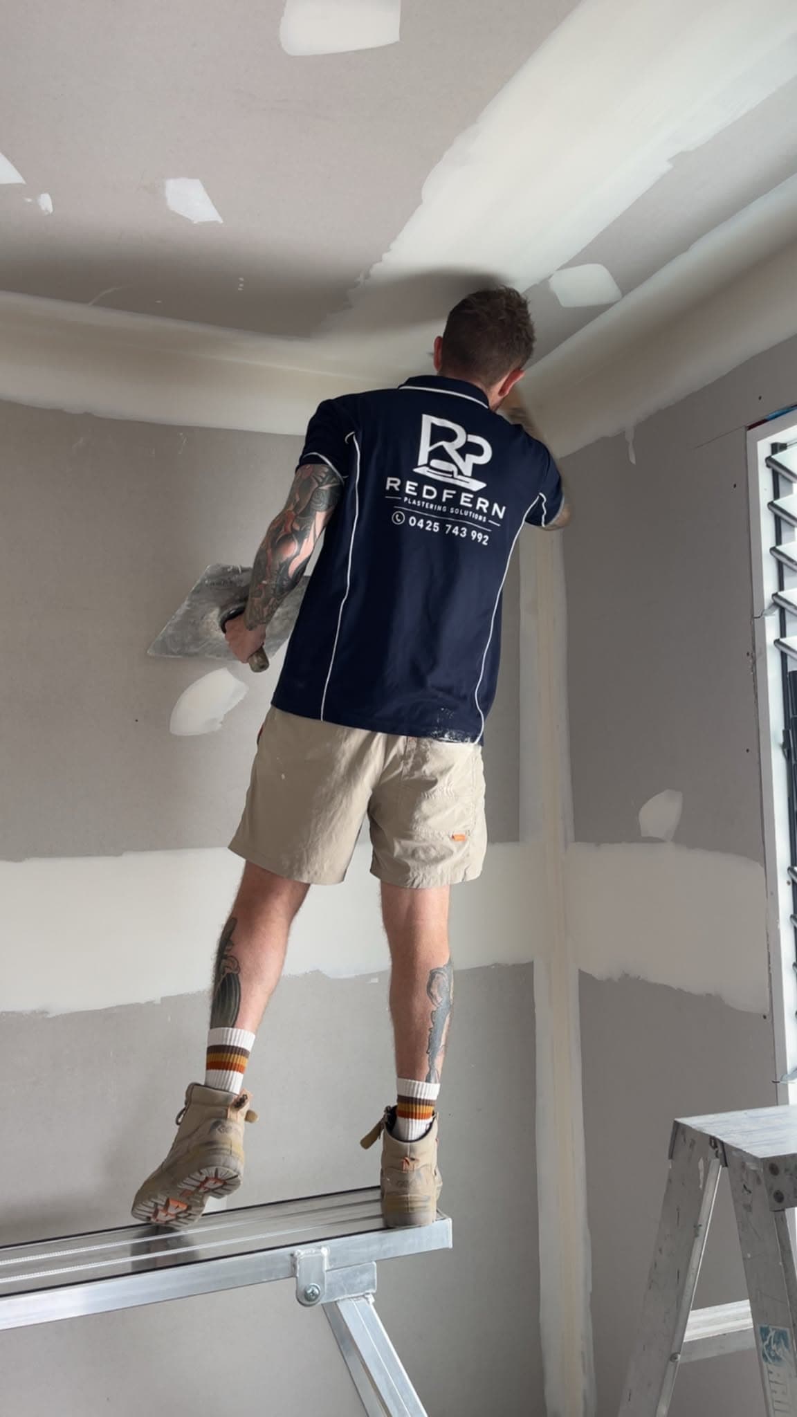 Plasterer on a platform ladder applying compound to a ceiling join in a Redfern branded shirt